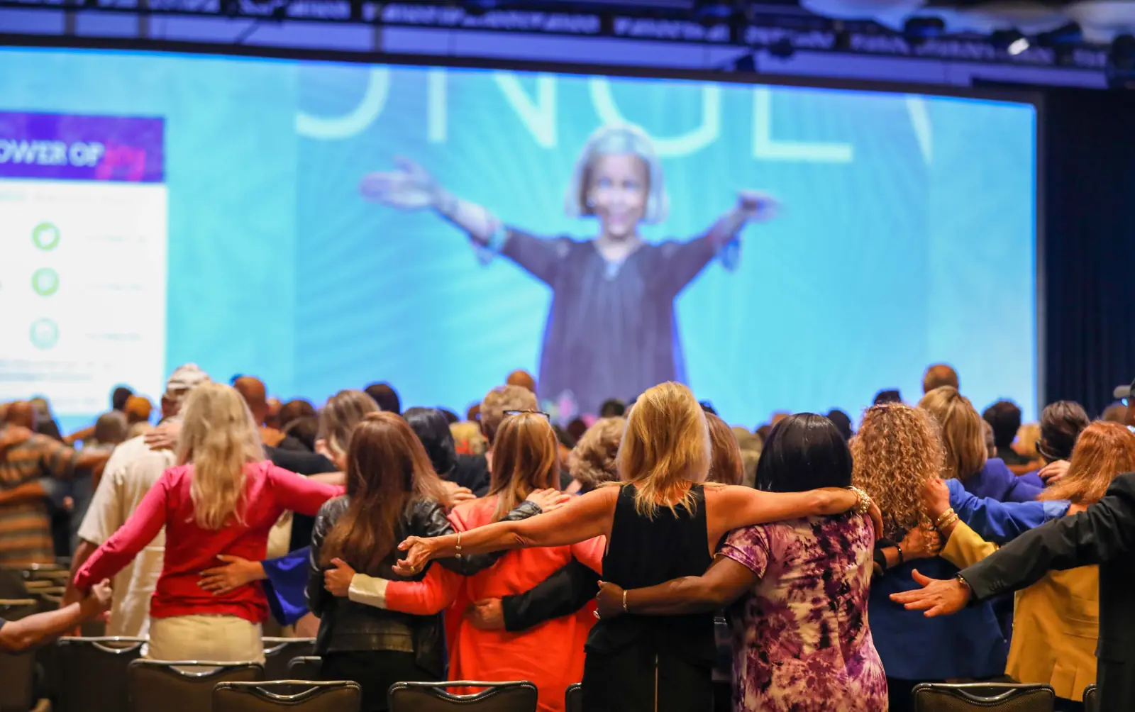 Amanda Gore, leadership keynote speaker, on the big screen with her arms wide while the audience stands with linked arms
