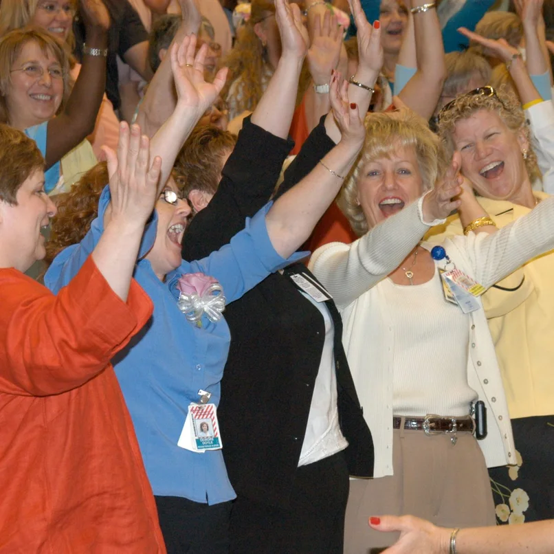 Women in the audience of an Amanda Gore keynote with their arms in the air, laughing