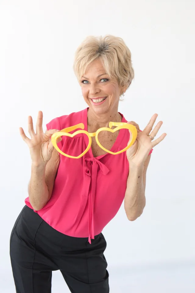 Amanda Gore, leadership keynote speaker, smiling, holding giant yellow heart-shaped glasses in front of her