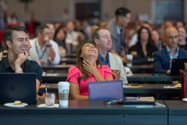 A woman in a pink dress laughing out loud with her head thrown back, in the audience at an Amanda Gore keynote