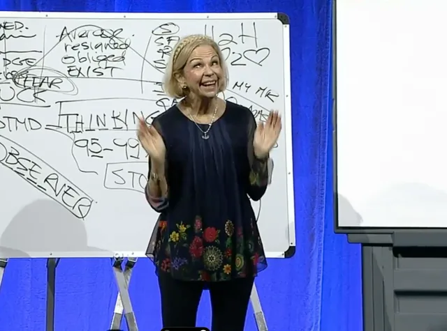 Amanda Gore, leadership speaker, on stage with both hands up, in front of a whiteboard covered in her notes