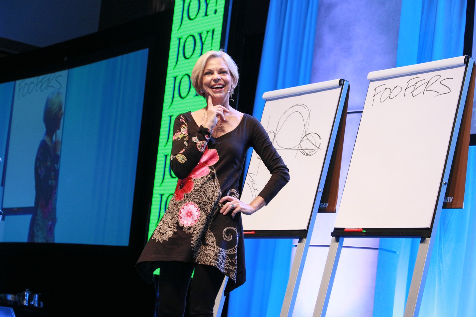 Amanda Gore, leadership keynote speaker, laughing on stage in front of a big JOY banner, one hand on her hip