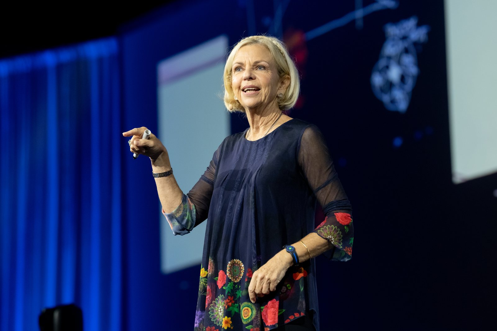 Amanda Gore, leadership keynote speaker, on stage pointing toward the audience in a navy top with a floral hem
