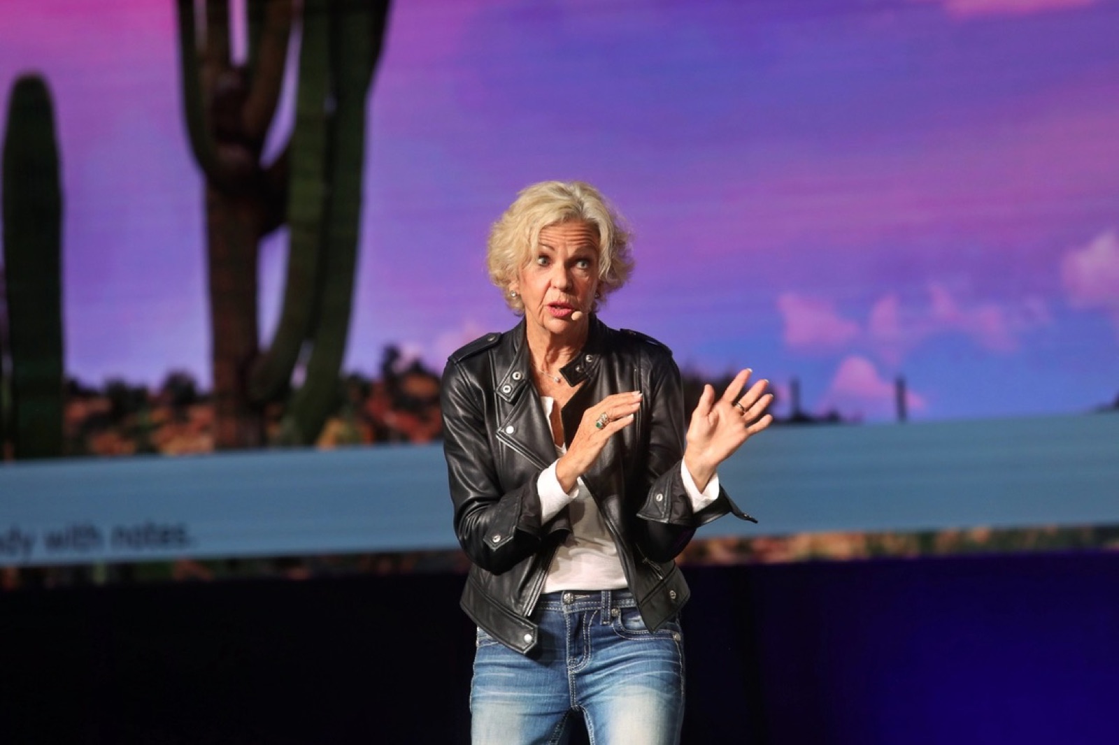 Amanda Gore, leadership keynote speaker, on stage in a leather jacket, speaking in front of a desert sunset screen