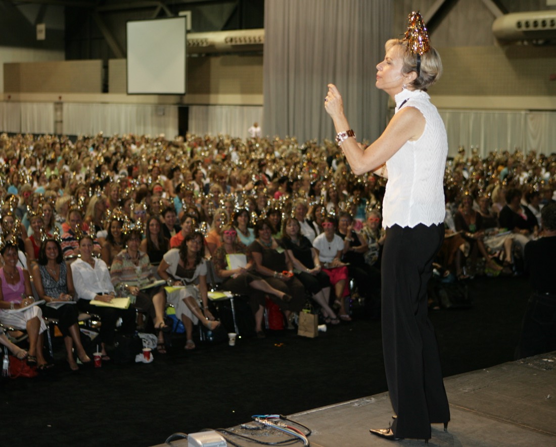 Amanda Gore, leadership keynote speaker, on stage wearing a gold tinsel headband, facing a packed room of women at a women’s leadership conference