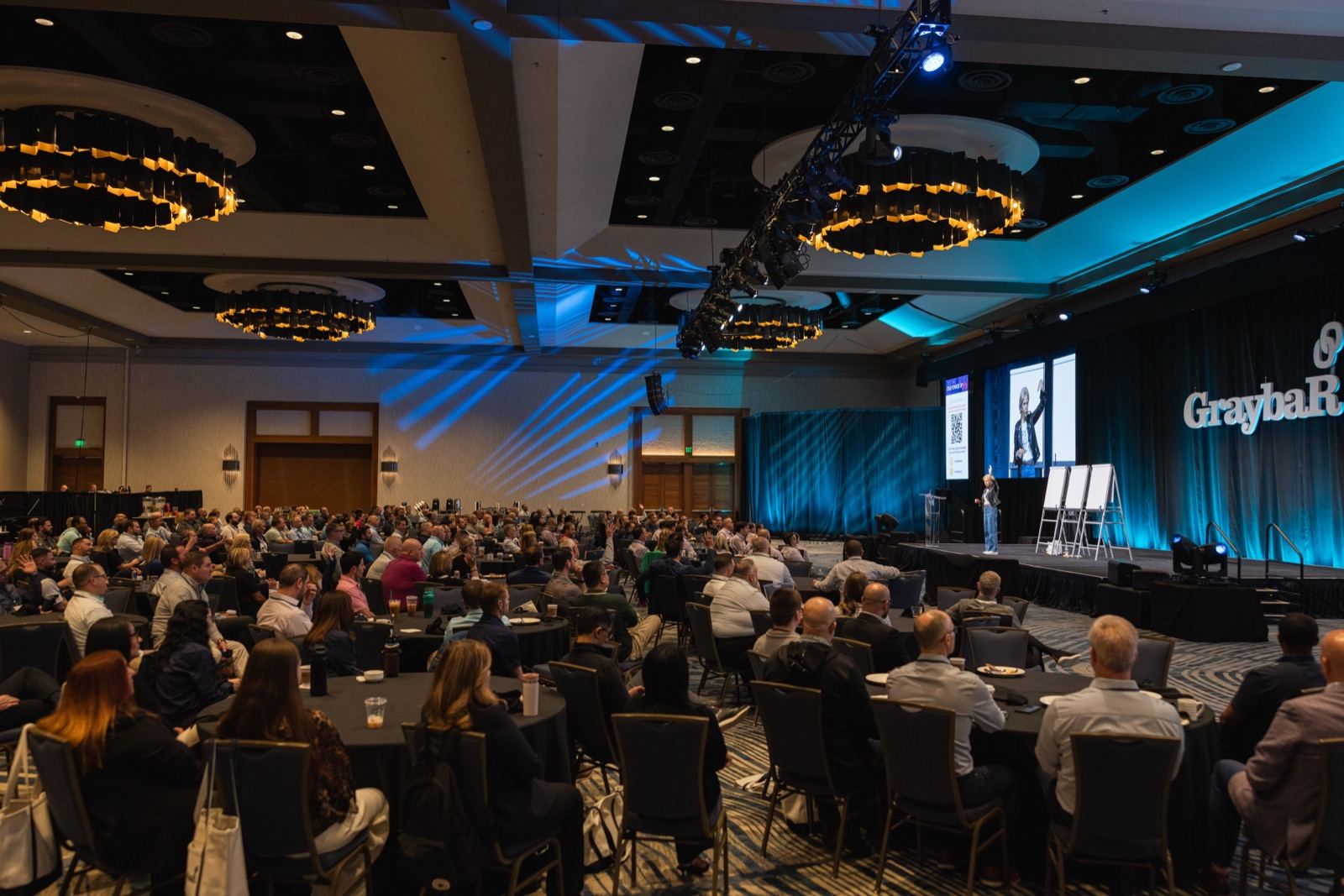 Amanda Gore, leadership keynote speaker, on stage between three flip charts at the Graybar conference, with a full ballroom in front of her