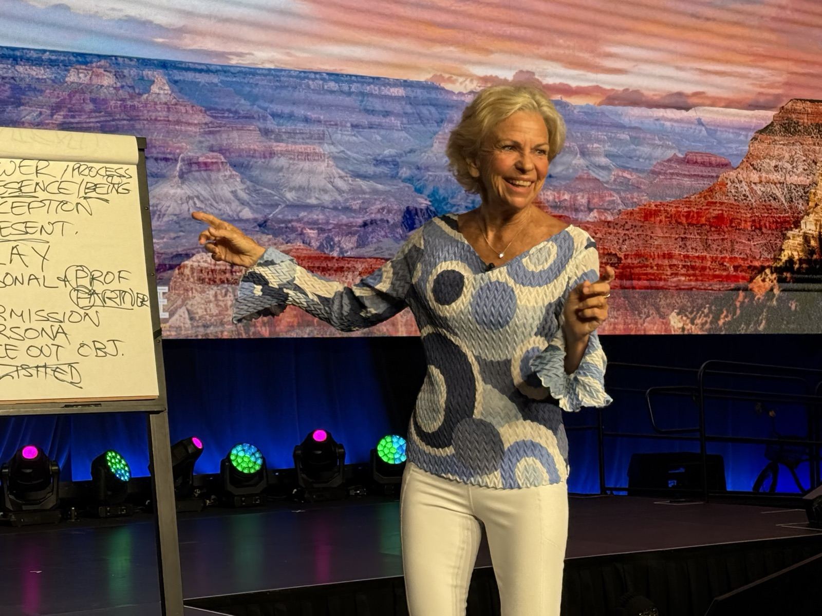Amanda Gore, leadership keynote speaker, laughing and pointing out to the audience on a conference stage, beside a flip chart